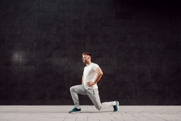 Side view of fit handsome muscular bearded focused sportsman in tracksuit doing lunges outdoors.