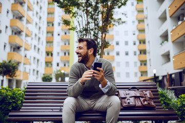 laughing handsome stylish businessman typing message on smart phone while sitting on the bench in park. Next to him is leather bag.