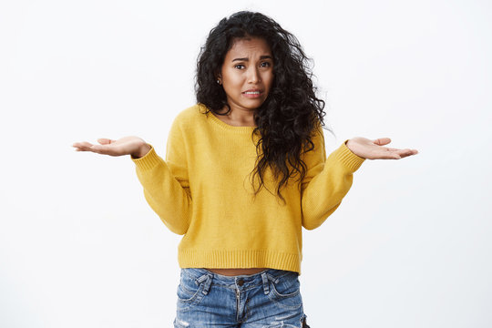 Oops girl says sorry. Cute silly african american woman, curly black hair, shrugging hands spread sideways, frowning and crying as feeling worried, apologizing making mistake, stand white background
