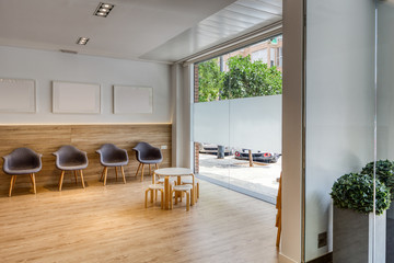 Modern design reception with gray chairs, pictures, white walls. Chairs and tables for children. Wooden floor and glass to the outside. Dental clinic.