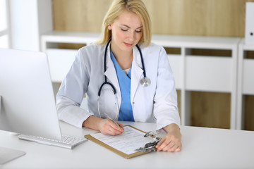 Doctor woman at work while sitting at the desk in hospital or clinic. Blonde cheerful physician filling up medical records form. Data and best service in medicine and healthcare