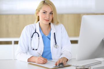 Doctor woman at work while sitting at the desk in hospital or clinic. Blonde cheerful physician filling up medical records form. Data and best service in medicine and healthcare