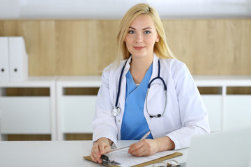 Doctor woman at work while sitting at the desk in hospital or clinic. Blonde cheerful physician filling up medical records form. Data and best service in medicine and healthcare