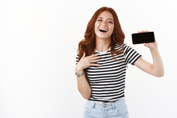Fototapeta premium Grateful and flattered cute redhead girl in striped t-shirt, showing smartphone display horizontally, touching chest thankful and touched, smiling happily, laughing and gazing camera