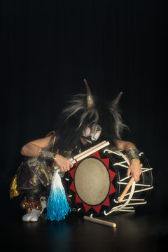 Taiko drummer in a wig and a demon mask on stage squarts with drumstick and drum and listen on a black background. Demon from Japanese mythology.