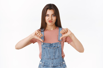 Disappointed skeptical arrogant attractive woman in overalls, t-shirt, smirking unsatisfied, showing thumbs-down and frowning in disagreement, express dislike judge something awful