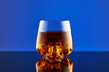 Crystal glass with a double portion of whiskey on ice on a glass surface and a blue background. A glass of whiskey is reflected on the surface.