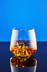Crystal glass with a double portion of whiskey on ice on a glass surface and a blue background. A glass of whiskey is reflected on the surface.