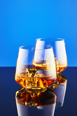 Two crystal glasses with a double portion of whiskey with ice on a glass surface and a blue background. The composition is reflected on the surface.