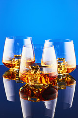 Three crystal glasses with a double shot of whiskey on ice on a glass surface and a blue background. The composition is reflected on the surface.
