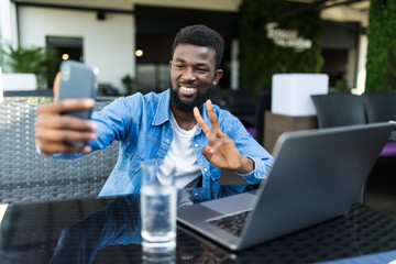 Young african man with phone take selfie make peace gesture while sitting with laptop in cafe