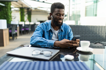 Portrait of happy african man using phone while working on laptop in a restaurant