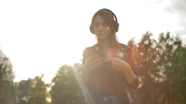 Portrait of a young woman puts on headphones and turns on the music on her smart phone. Music lover concept. A female in a leather jacket walks in the park. Slow motion. Sunset orange flare.