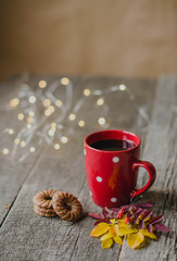 Autumn still life. Red Cup, cookies, colored leaves on a wooden rustic table. In the background the lights of garland. Top view from the side.