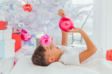 Up-view shot of little child laying under christmas tree, touching balls, looking at his reflection in ornaments glass.