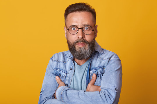 Portrait of shocked middle aged man in full disbelief. Hipster male with beard wears denim jacket posing isolated over yelow studio background. Astonished face expression, human emotion, body language