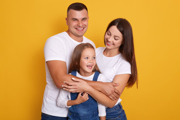 Happy family looking very excited, posing isolated over yellow background, mother, father and daughter wearing white shirts, woman and man hugging charming girl with great love. Happyness concept.