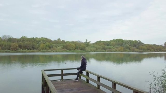 A lonely woman sits warmly dressed on a jetty by a lake in autumn.The camera accelerates quickly over her head and over the water. Overcast weather and smooth reflecting seawater. A duck on the lake. 