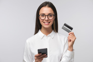 Young smiling woman holding credit card and phone, isolated on gray background