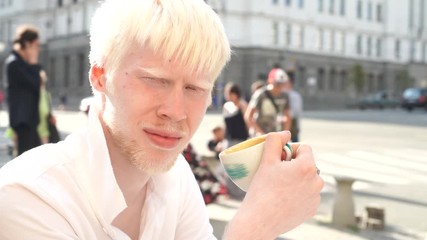 portrait of an albino man outdoor dressed t-shirt sitting outdoor cafe and drink coffee. abnormal deviations. unusual appearance