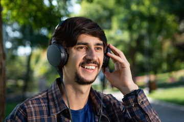 Handsome brunet bearded man in a lumberjack shirt listening to music enjoying warm day in a park.