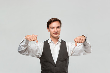 Handsome man in a white shirt and grey vest showing with his fingers down at the empty space for advertising.
