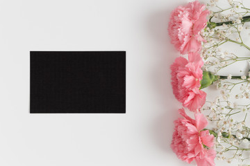 Top view of a black card mockup with gypsophila and pink carnations on a white table.