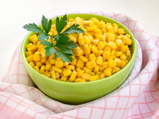 Close-up of canned sweet corn in a green bowl near checkered kitchen towel. Garnish and ingredient for salads. Vegetarian.