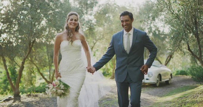 Happy multi ethnic bride and groom joyfully running together down a path on their wedding day in nature, diverse couple holding hands and eloping together on their wedding day