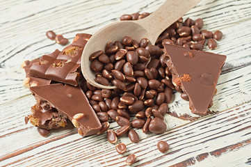 Many dragees with glazed peanuts strewn from a wooden spoon near broken chocolate bar with caramel, raisin, big nuts on old worn white rustic table. Close-up