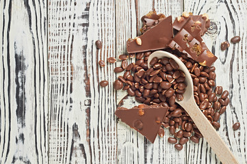 Lot of dragees with glazed peanuts strewn from a wooden spoon near broken chocolate bar with caramel, raisin, big nuts on old worn white rustic table. Top view. Space for text