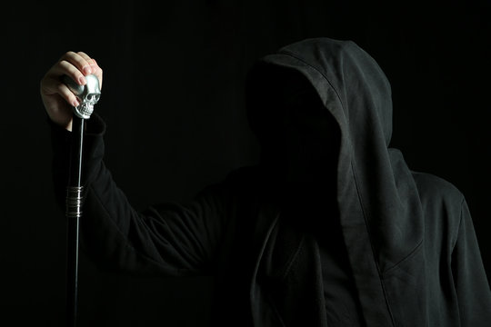 Man Without Face In A Black Hood Hold A Cane With A Skull On Dark Background