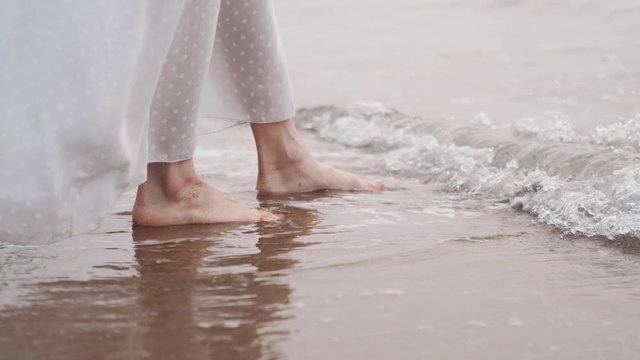 Female feet on the seashore, touch the sand, water. Dress develops in the wind.