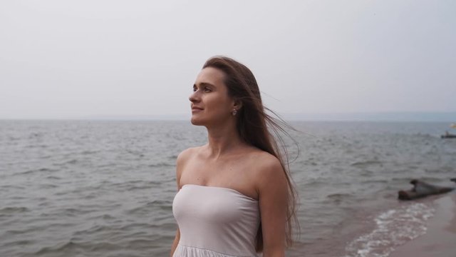 An attractive girl in a white dress walks along the seashore, the wind develops her long hair.