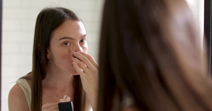 Woman puts foundation on her face while looking in reflection in a bathroom mirror