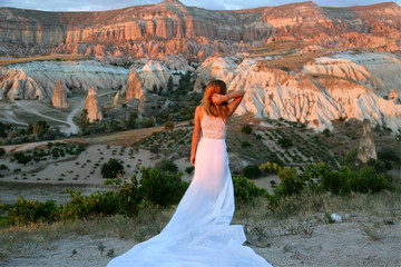Happy  young woman traveller in long  white dress stands on a mountain against the background of balloons in Cappadocia Turkey. Visit Turkey. Amazing view.