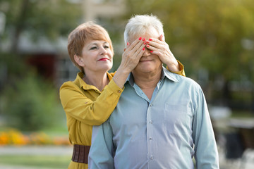 Elderly married couple outdoors. Outdoors in the park A woman of age gently covers her retiree's eyes with her husband.