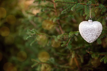 Close-up of Christmas festive decoration toy shaped like a heart hanging on a fir tree branch with bokeh blured lights. Green wood background. Copy space.