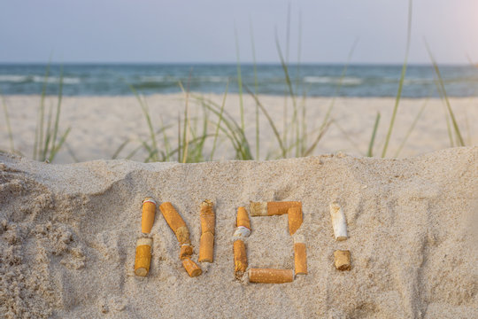 in the foreground, the word "no!" written in the  sand with cigarette stubs,  the sea is in the background
