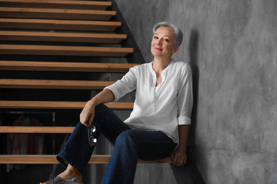 Happy mature woman sitting on wooden stairs indoors. Smart aging