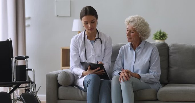 Young woman doctor help disabled old patient write in clipboard