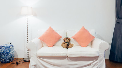 Two seater sofa in living room with bear, lamp and blank artwork.White Couch in an Architectural White Living Room, with white Wall, Decorated with Standing Lampshade and teddy bear