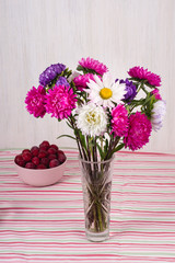 Red plums in pink bowl and glass vase with a bouquet of chrysanthemums on  colorful striped tablecloth.