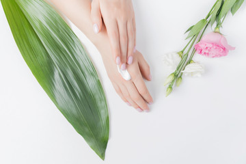 Girl puts cream on hands on white background with tropical leaves and pink flowers, top view. Concept of skin care, anti-aging frost
