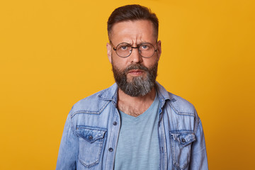 Obraz premium Close up portrait of beautiful bearded guy with stylish hairstyle looks directly at camera with serious and calm facial expression, poses isolated on yellow studio background. People emotions concept.
