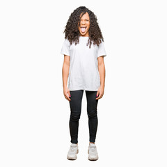 Young beautiful woman with curly hair wearing white t-shirt sticking tongue out happy with funny expression. Emotion concept.