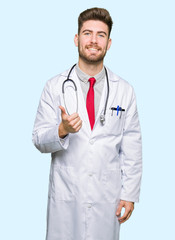 Young handsome doctor man wearing medical coat doing happy thumbs up gesture with hand. Approving expression looking at the camera showing success.