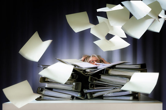 desperate woman sinks behind stacks of ring binders on an office desk and lots of papers are flying down, concept of excessive demands and increasing work in business
