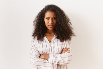 Unhappy stubborn young African American female in striped shirt posing isolated in closed posture, keeping arms crossed, frowning, expressing dissatisfaction or disagreement, feeling offended - obrazy, fototapety, plakaty