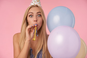 Attractive young blonde female with casual hairstyle wearing blue cone hat and blowing party horn, celebrating birthday with multicolored air balloons, having cheerful moments in her life during party